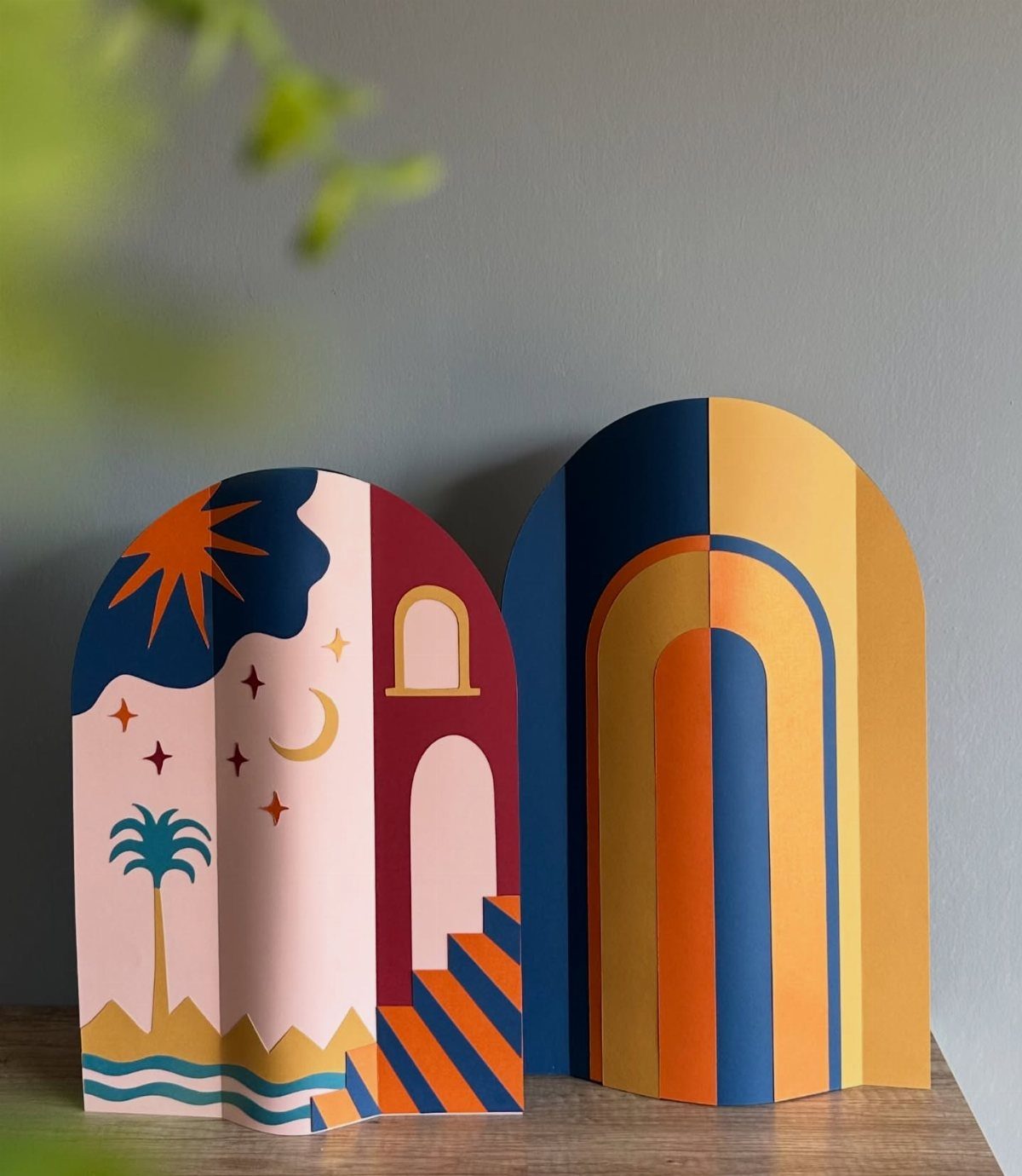 23 DIY Decor with Paper Projects for Every Room 16 Two arched folded paper sculptures with colorful desert and rainbow designs on wood.