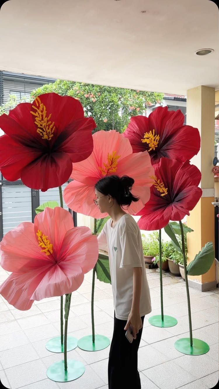 23 DIY Decor with Paper Projects for Every Room 20 Giant red and pink paper hibiscus flowers standing on green stems beside a woman.
