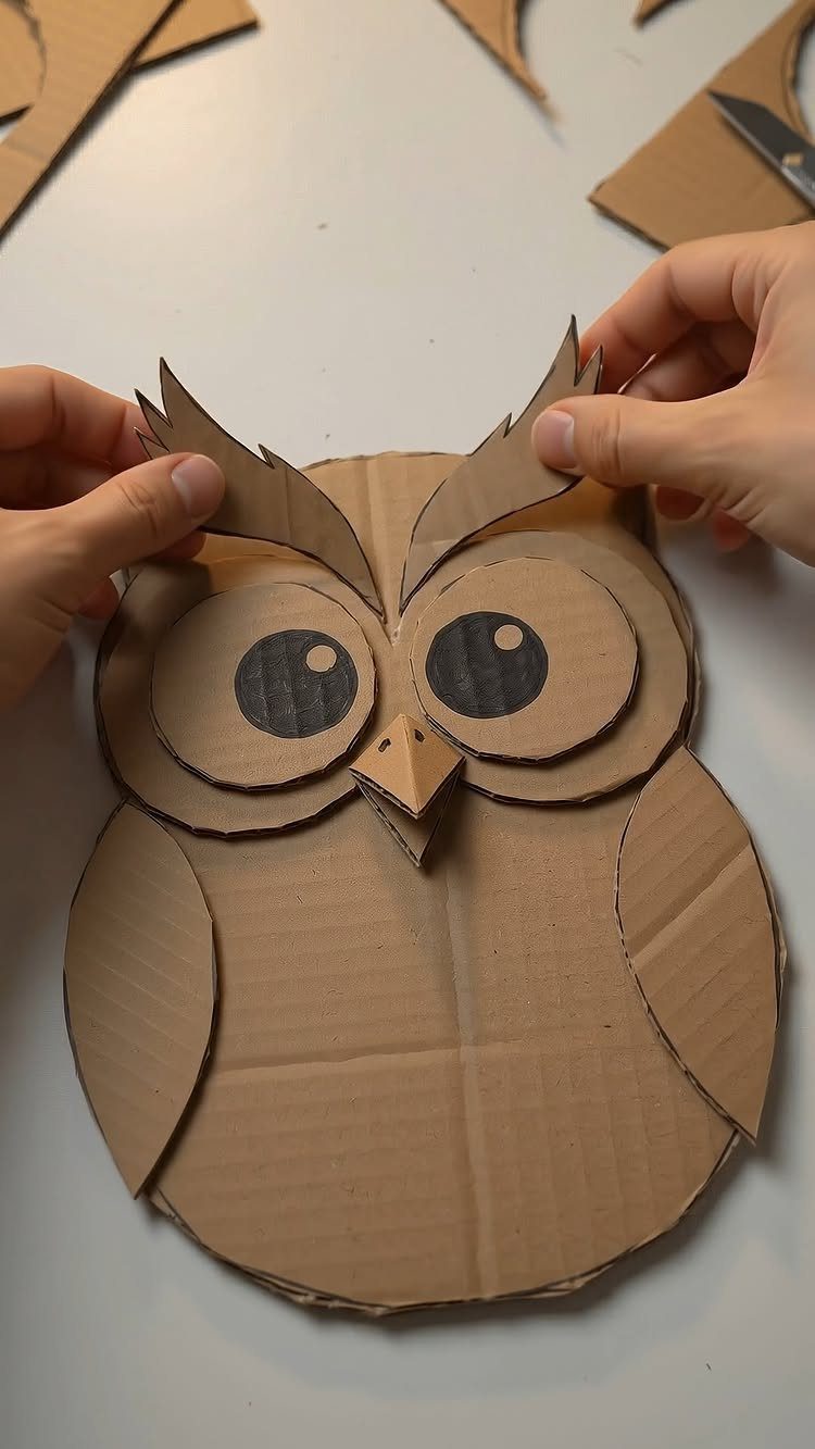 17 Recycled Art Projects for Kids and Adults to Make 16 Cardboard owl figure being assembled by hands on a white tabletop surface.
