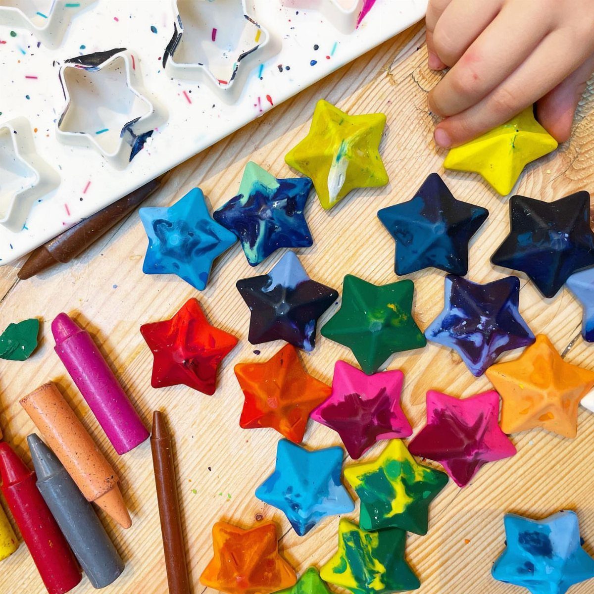 17 Recycled Art Projects for Kids and Adults to Make 10 Star-shaped colorful crayons on a wooden table with a child's hand and molds.