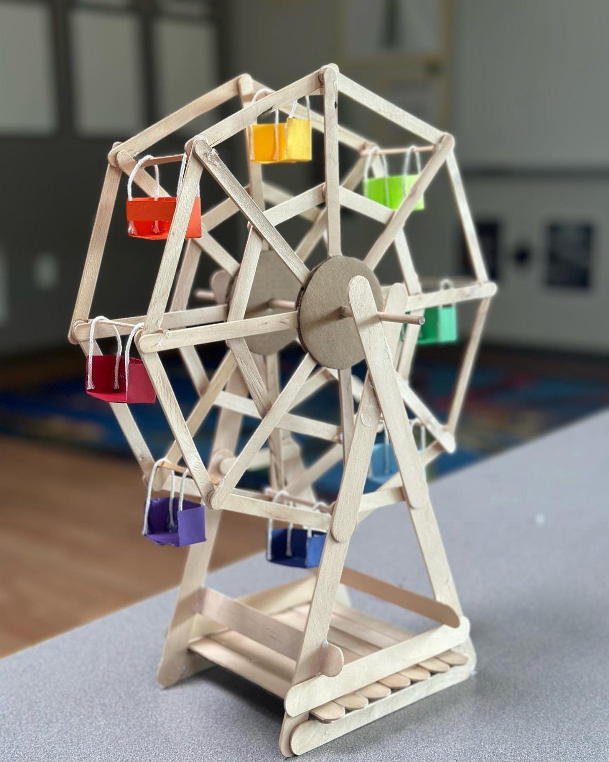 17 Recycled Art Projects for Kids and Adults to Make 12 Popsicle stick Ferris wheel model with colorful paper gondolas on a grey surface.